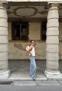 Ανοιχτό μπεζ πλεκτό τοπ, ανοιχτό μπλε παντελόνι με φαρδιές γραμμές και μια λουλουδάτη τσάντα πλάτης. Στέκεται ανάμεσα σε δύο πέτρινες κολώνες με υφές στους τοίχους πίσω.