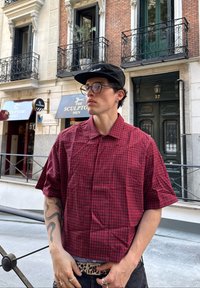 Camicia a maniche corte a scacchi rossi e neri con colletto strutturato. Indossata sopra jeans scuri, abbinata a una cintura con stampa leopardata.