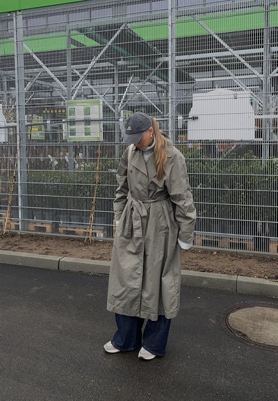 Un abrigo trench largo de color gris claro con cintura ajustada por un cinturón, botones grandes y mangas extragrandes, combinado con unos vaqueros anchos azules y zapatillas blancas.