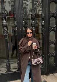 Femme en manteau marron et lunettes de soleil tenant une tasse de café et un sac à main à motifs, debout devant des portes en métal noir orné et en verre.