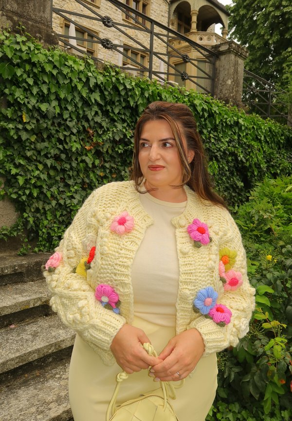 Kvinde iført en cremefarvet sweater med farverige strikkede blomster, der holder en cremefarvet håndtaske, stående foran stentrapper og en grøn mur med vedbend.