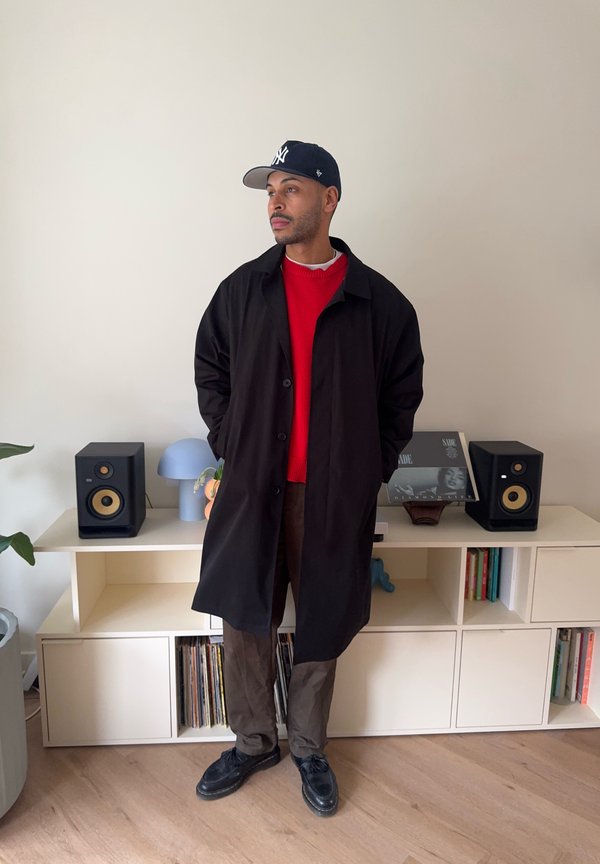 Mand iført sort frakke, rød sweater og mørke bukser står foran hvide reoler med højtalere og bøger i en moderne stue.