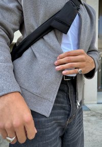 Egy szürke dzsekit, fehér inget, fekete farmernadrágot és gyűrűket viselő személy, aki az egyik kezével a dzsekit igazítja, míg a másik kezében egy fekete keresztpántos táskát hord.