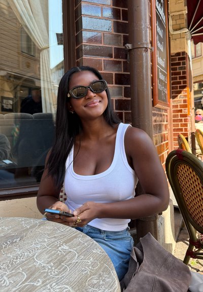 Una mujer con una camiseta blanca está sentada en una mesa de café, sosteniendo un teléfono inteligente. Lleva gafas de sol negras y tiene el cabello largo y lacio.