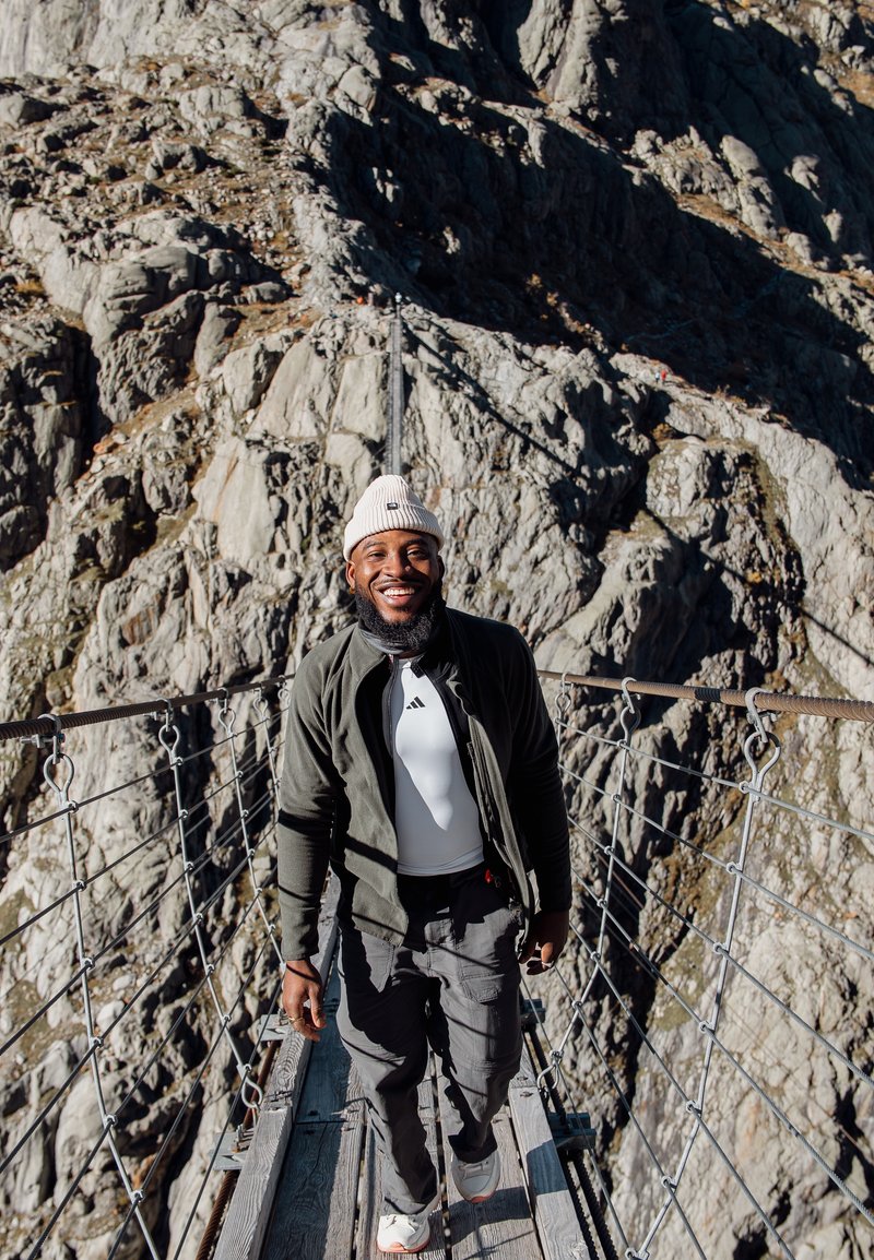 A person on a suspension bridge, wearing a cream beanie, a black zip-up jacket over a white shirt, grey cargo trousers, and trainers. A rocky backdrop.