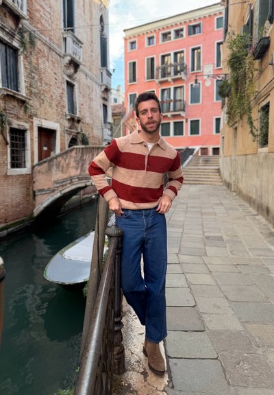 Hombre con suéter a rayas rojo y beige y vaqueros azules apoyado en una barandilla junto a un canal con un barco, edificios históricos y camino de piedra de fondo.