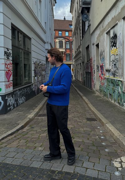 Suéter de punto azul acanalado, pantalones negros holgados y zapatos negros gruesos; de pie en un callejón estrecho cubierto de grafitis con pavimento de adoquines.