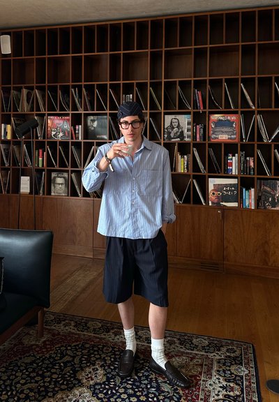 Camisa de botones a rayas azul claro, pantalones cortos a rayas azul marino, calcetas blancas, mocasines oscuros, diadema negra. El fondo presenta estanterías de madera con libros y discos.