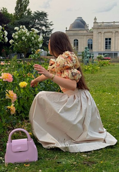 Una persona se arrodilla en un jardín con una blusa floral de mangas abullonadas y una falda larga de un tono beige claro. Un pequeño bolso rosa descansa en la hierba cercana.