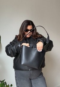 Mustast nahast käekott kõverate õlarihmade ja minimalistliku disainiga, mida hoiab inimene, kes kannab musta üle suuruses jope.