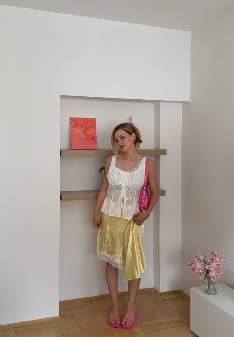 Femme debout à l'intérieur portant un haut en dentelle blanche, une jupe en satin jaune, des sandales roses, et tenant un sac rose, à côté d'étagères en bois et de fleurs.