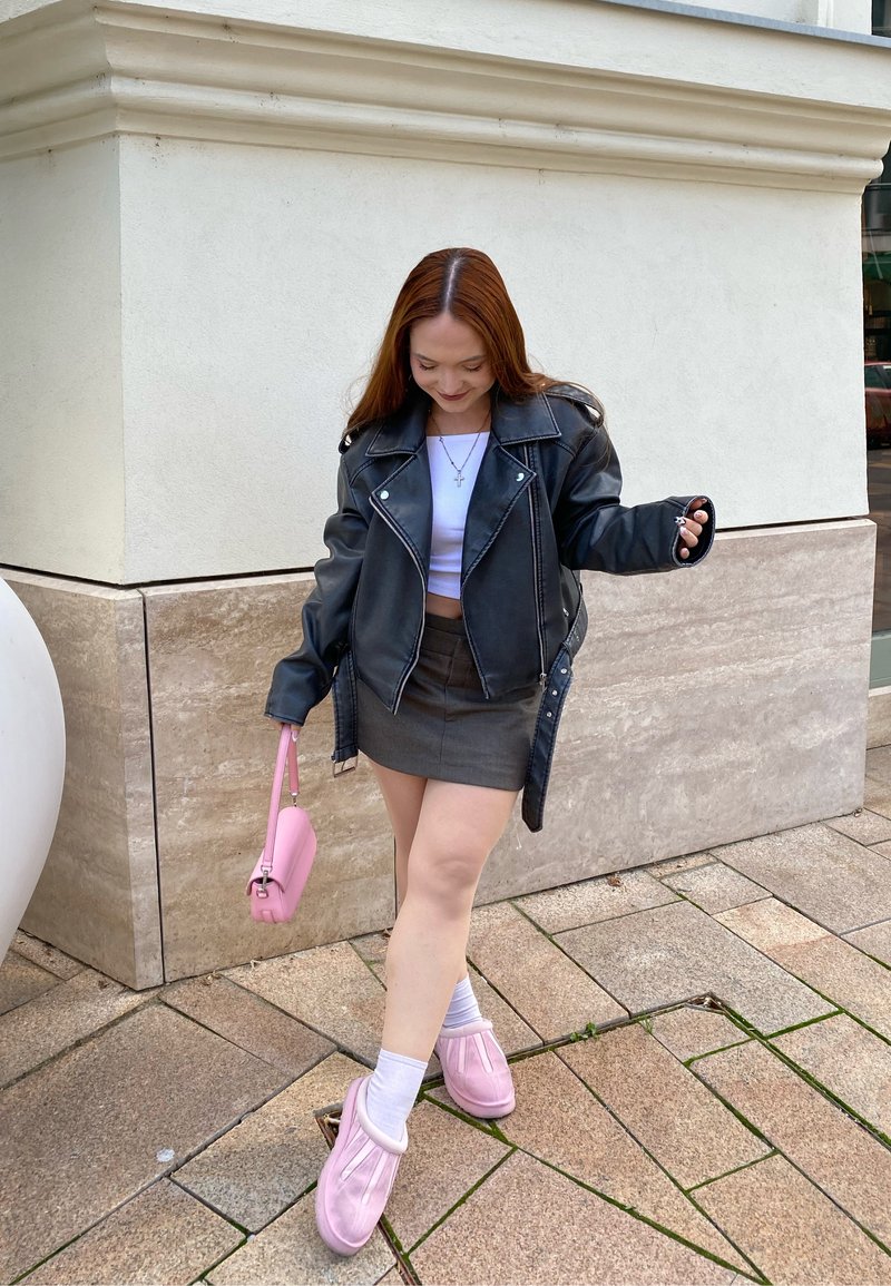 Schwarze Leder-Motorradjacke, weißes Cropped-Top, grauer Minirock, pinke Handtasche, pinke flauschige Hausschuhe, stehend auf Steinpflaster.
