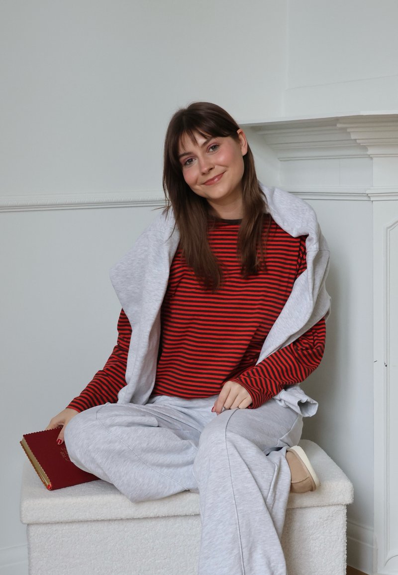 Jeune femme aux longs cheveux bruns assise en tailleur sur un banc blanc, portant un t-shirt rayé rouge et noir et un pantalon gris clair, tenant un livre rouge.