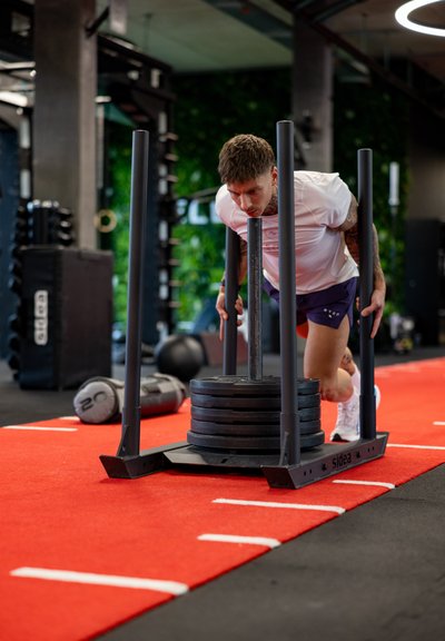 Hombre empujando un trineo con peso sobre un suelo de gimnasio rojo, vistiendo una camiseta blanca y pantalones cortos azules, concentrado en un entrenamiento de fuerza intenso.