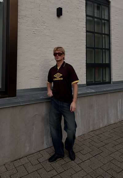 Camisa marrón de manga corta con detalles amarillos y logo. Jeans de mezclilla oscura y zapatos negros. De pie contra una pared texturizada de color claro.