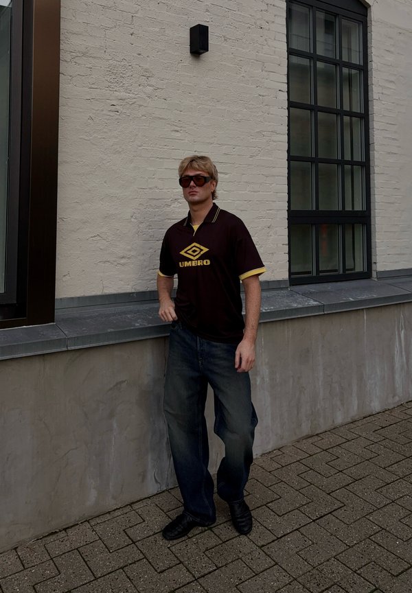 Pruun lühikeste varrukatega särk kollaste aktsentidega ja logo. Tumedad teksad ja mustad kingad. Seisab tekstureeritud heledal seinal.