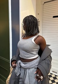 Persona ar pītām matiem, valkājot brilles, pelēku bezroku topu, vaļīgas pelēkas bikses ar džempera mezglu ap vidukli un tumši pelēku mēteļu, stāv iekštelpās.