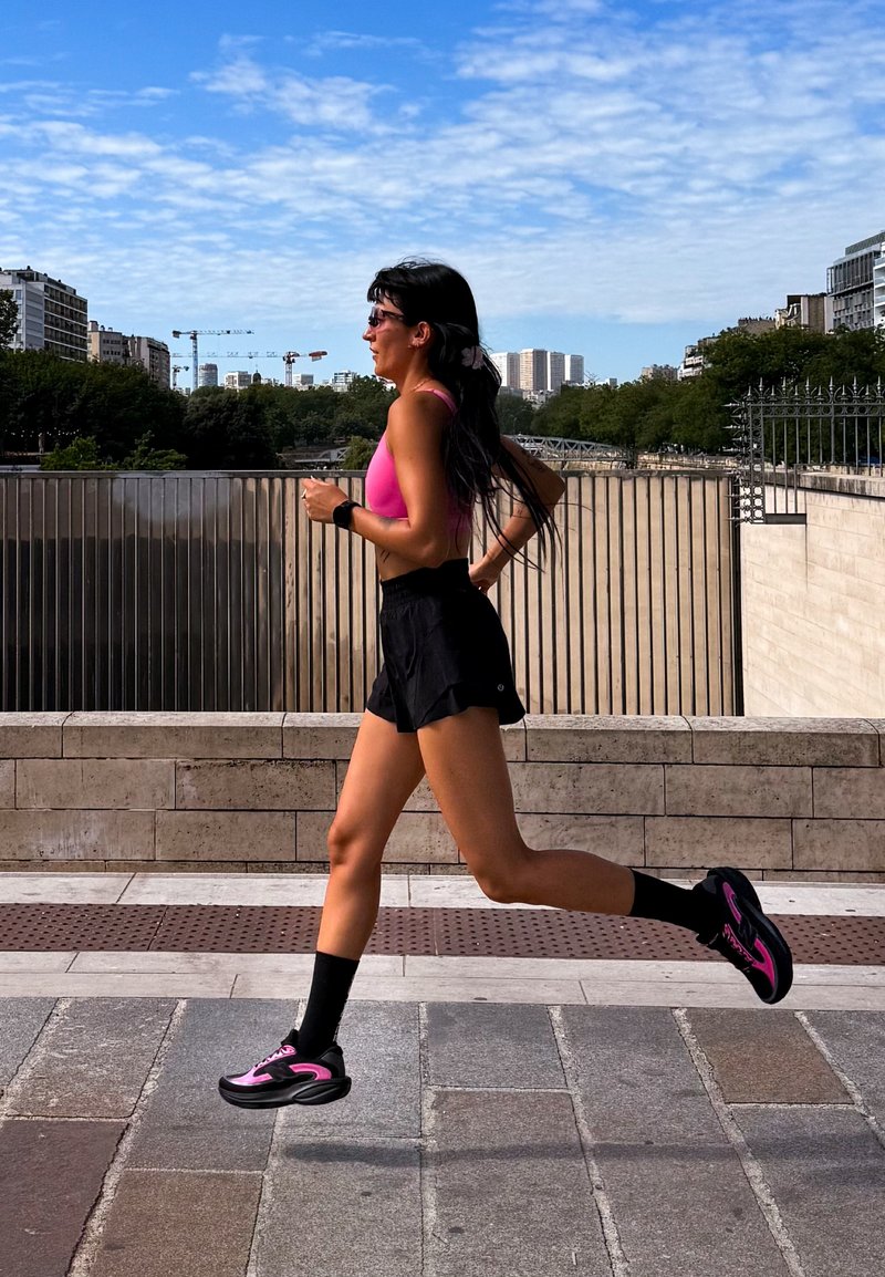 Femme faisant du jogging en plein air sur un chemin pavé, portant un haut de sport rose, un short noir et des chaussures de course, avec la ligne d'horizon de la ville et des arbres en arrière-plan.