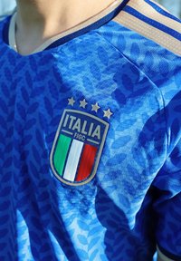 Μπλε εθνική φανέλα ποδοσφαίρου της Ιταλίας με το σήμα της Ιταλίας, που διαθέτει τέσσερα χρυσά αστέρια και πράσινες, λευκές, κόκκινες ρίγες στο στήθος.