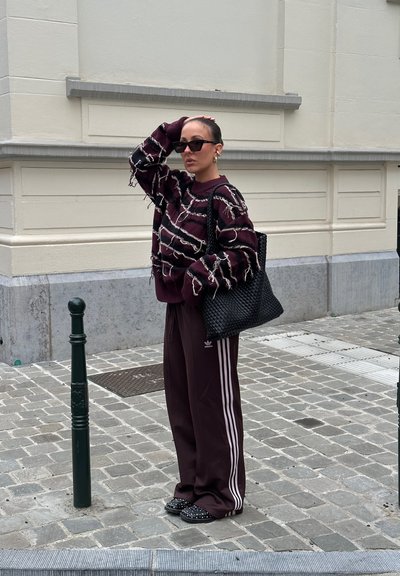 Suéter burdeos con detalles de flecos, pantalones de chándal amplios burdeos con franjas blancas, bolsa de mano tejida negra y zapatos sin cordones negros con tachuelas.