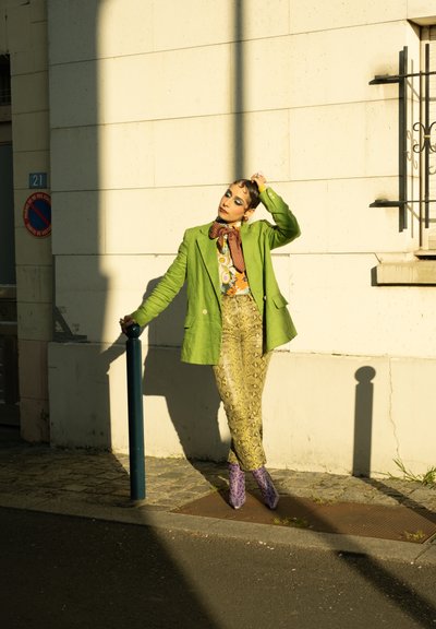 Blazer de lino verde sobre una camisa naranja estampada, combinado con pantalones de serpiente y botas de tacón moradas. La luz del sol proyecta sombras intensas.