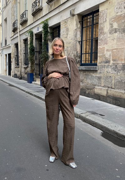 Conjunto de dos piezas de color marrón arrugado, con mangas largas, pantalones de pierna ancha y una blusa peplum de capas; acompañado de zapatos plateados y un bolso ligero.
