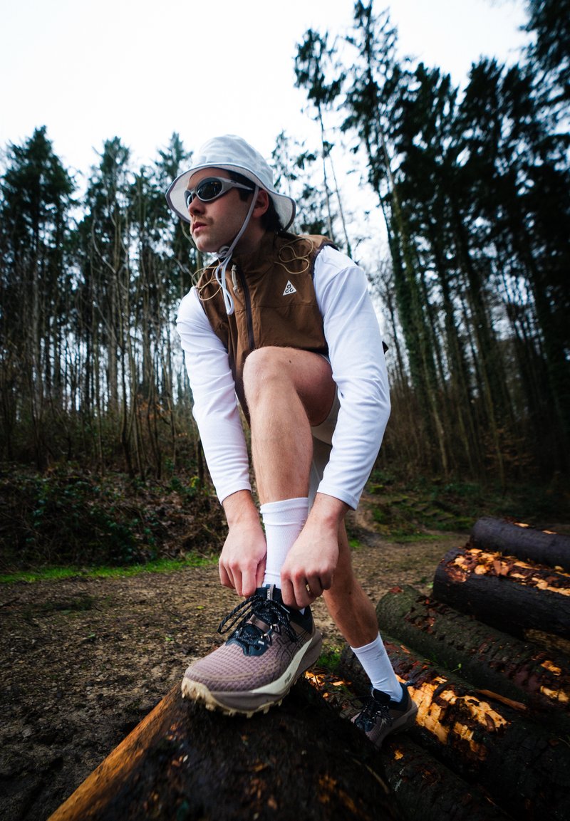 Άνδρας με γυαλιά ηλίου, λευκό καπέλο και καφέ γιλέκο δένει το παπούτσι του πάνω σε κορμό σε μονοπάτι δάσους με ψηλά δέντρα και συννεφιασμένο ουρανό.