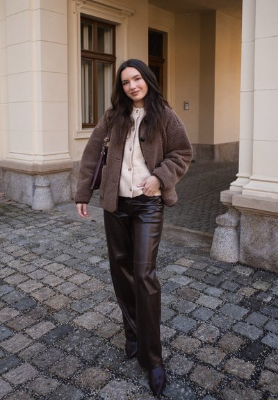 Joven mujer con chaqueta de peluche marrón, suéter beige, pantalones de cuero oscuro y botas, de pie en una calle adoquinada junto a un arco de edificio.