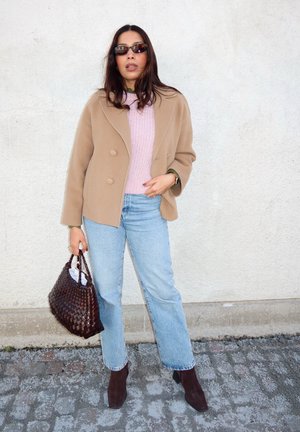 Mujer con abrigo color topo, suéter rosa, jeans azul claro, gafas de sol y botas marrones, sosteniendo un bolso tejido marrón frente a una pared lisa.