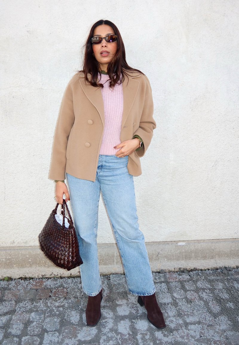 Mujer con abrigo color topo, suéter rosa, jeans azul claro, gafas de sol y botas marrones, sosteniendo un bolso tejido marrón frente a una pared lisa.