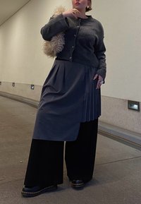 Grau gestrickte Wolle-Cardigan mit strukturierten Knöpfen, über einem gefalteten dunklen Rock und schwarzen weit geschnittenen Hosen getragen. Klobige schwarze Schuhe vervollständigen das Outfit.
