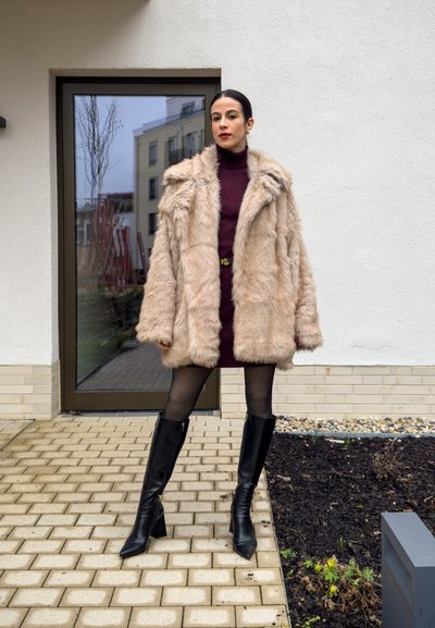 Abrigo de piel sintética en beige claro con un diseño texturizado, llevado sobre un vestido burdeos. Botas negras hasta la rodilla complementan el atuendo.