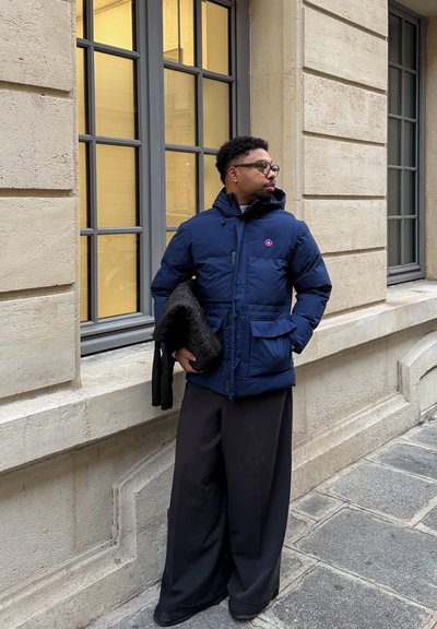 Chaqueta acolchada azul marino con capucha, cierre de cremallera y bolsillos delanteros. Se lleva sobre pantalones negros de pierna ancha, sujetado con una prenda acolchada negra.