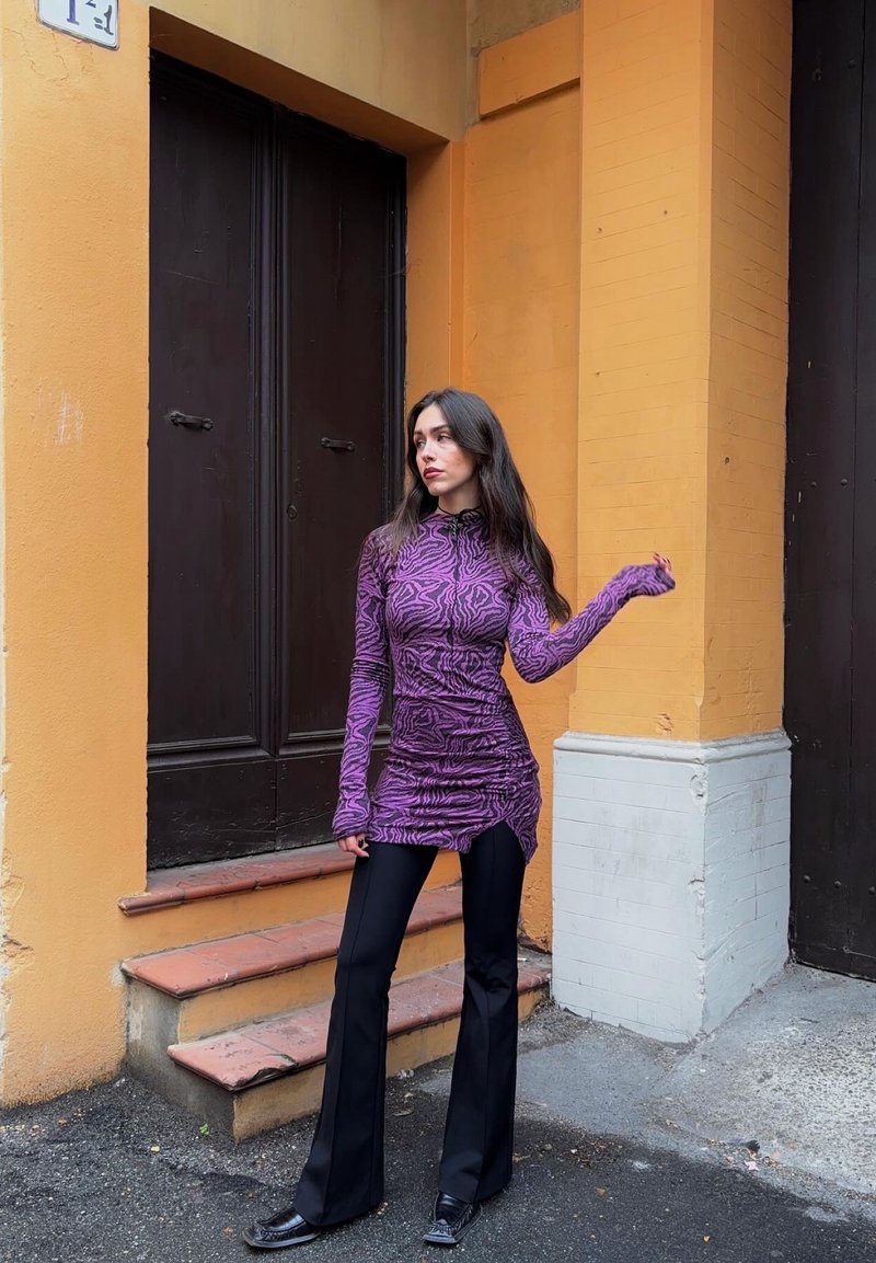 Haut violet à manches longues avec un motif ondulé et des côtés froncés, associé à un pantalon noir ajusté évasé et des chaussures noires pointues.