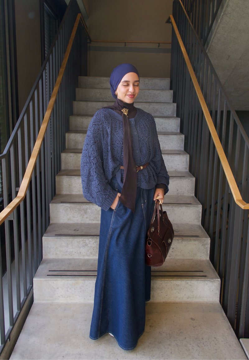 Gestrickter blauer Pullover mit komplexen Mustern, weiten Ärmeln, kombiniert mit einem langen Jeansrock. Eine braune Handtasche vervollständigt das Outfit. Stehend auf einer Treppe.