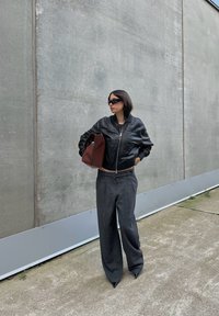 Svart läder bomberjacka, grå vida skräddarsydda byxor och spetsiga svarta klackar. Håller en brun väska, mot en grå vägg.