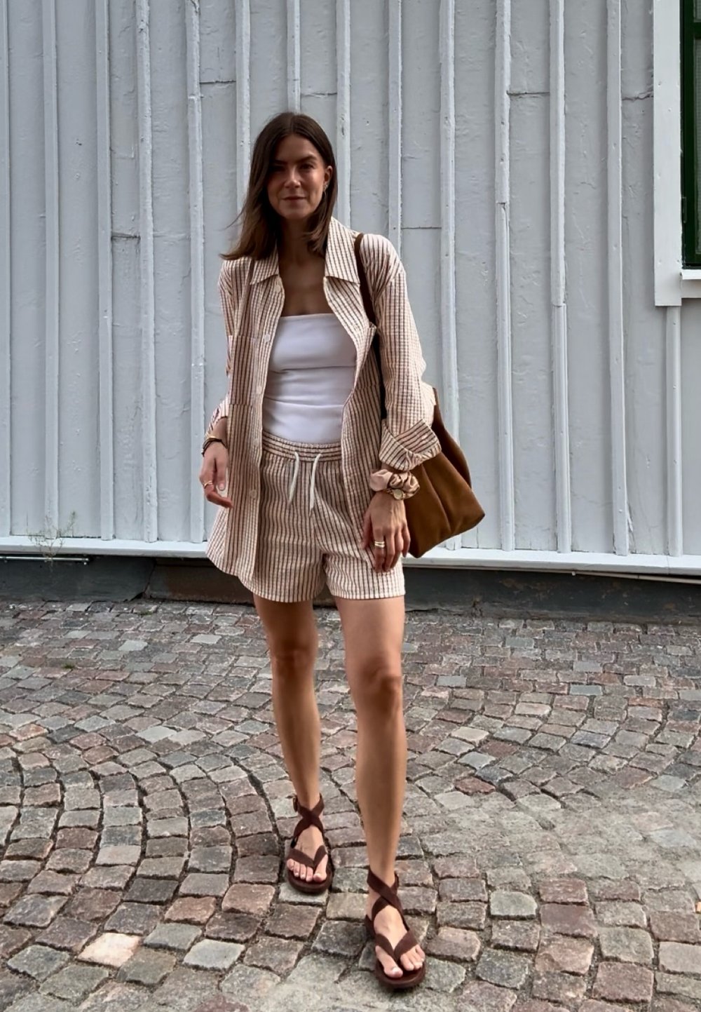 Nő sétál a macskaköves utcán bézs csíkos kabátban és rövidnadrágban, fehér felsőt visel, barna szandál van rajta, és barna válltáskát hord.