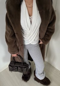 Manteau en fausse fourrure marron, haut blanc torsadé, pantalon de survêtement gris et bottes doublées de fourrure. Tient un sac marron foncé avec deux poches frontales.