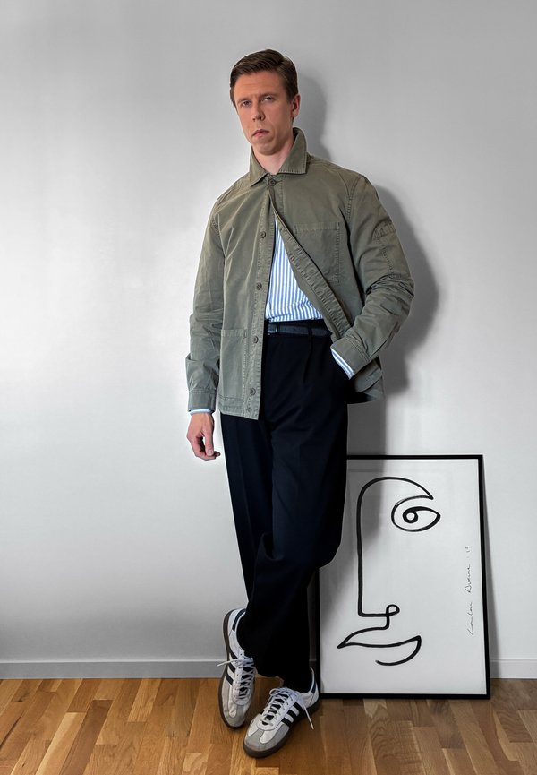 Homme en veste verte, chemise rayée, pantalon noir et baskets appuyé contre un mur blanc à côté d’un dessin abstrait de visage encadré sur un sol en bois.