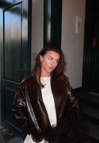 Hnědá lesklá bomber bunda se zipem, nošená přes bílý top, doplněná černým texturovaným doplňkem. Jednobarevné zdi a dveře.