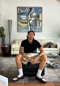 Man met tatoeages in een zwart shirt en beige shorts zittend op een zwarte ottoman in een woonkamer met een abstract schilderij en een witte bank achter hem.