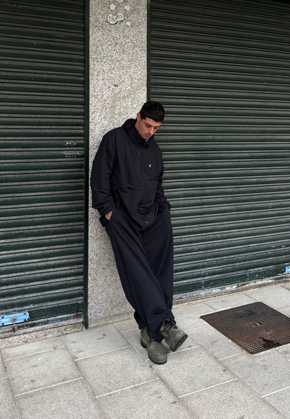 Chaqueta negra con capucha y un bolsillo frontal, combinada con pantalones negros holgados y botas oscuras, de pie frente a una pared texturizada.