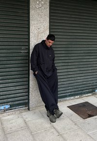 Chaqueta negra con capucha y un bolsillo frontal, combinada con pantalones negros holgados y botas oscuras, de pie frente a una pared texturizada.