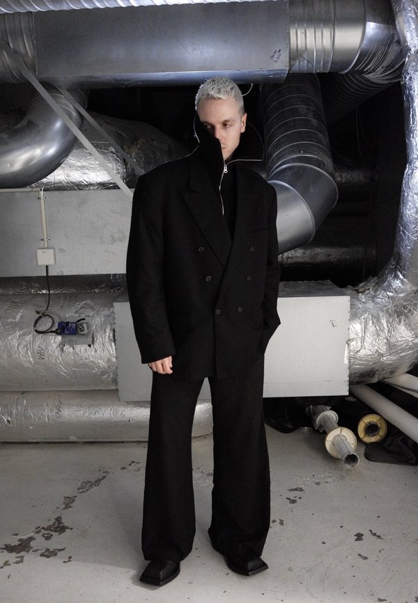 Personne aux cheveux blonds courts portant une veste et un pantalon noirs surdimensionnés, debout dans un espace industriel avec des tuyaux et des conduits métalliques.