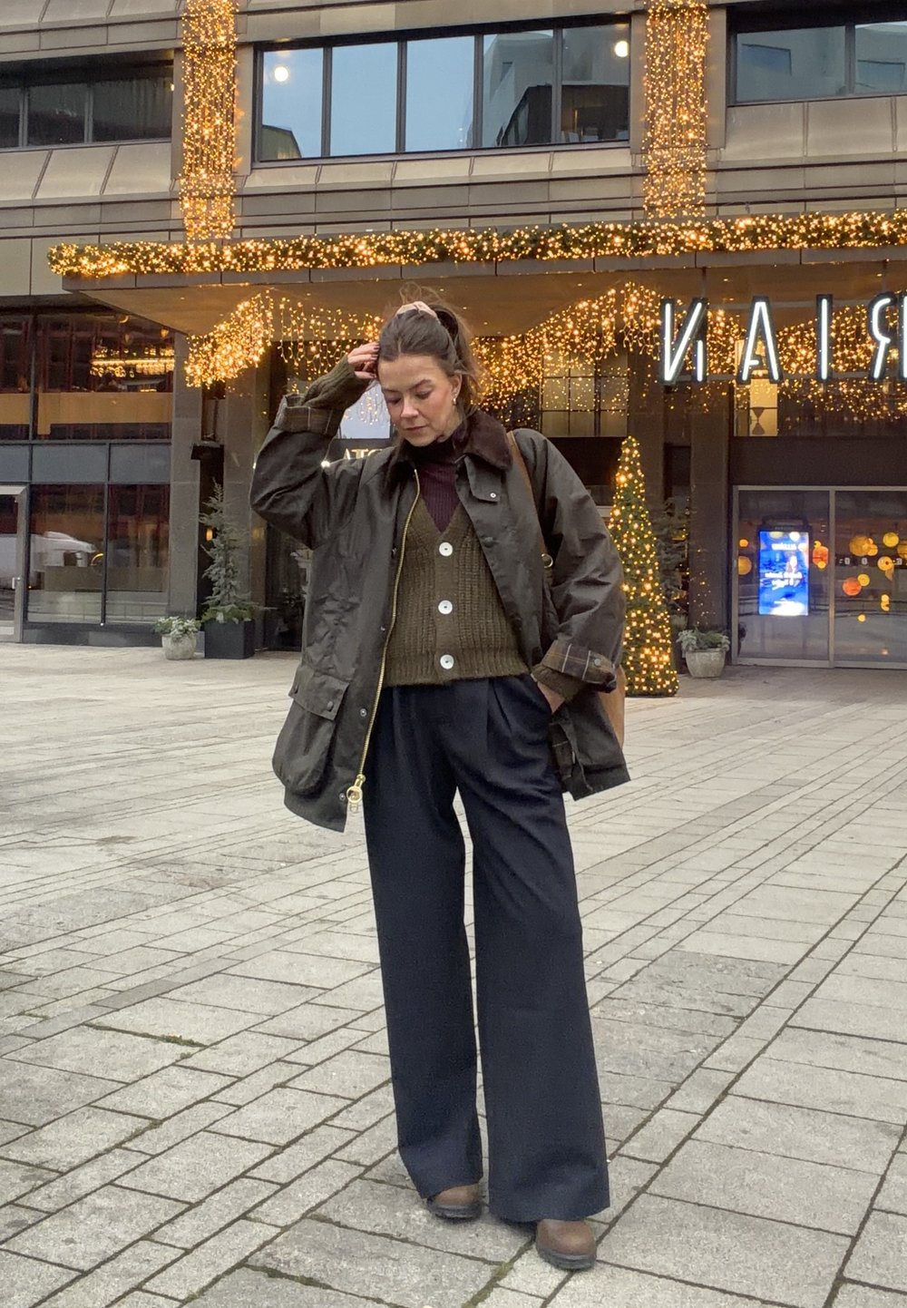 Donkergroene waterdichte jas met een bruine kraag, een olijfgroene trui en brede zwarte broek. De outfit heeft oversized knopen en een casual pasvorm.