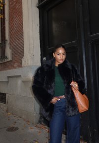 Musta keinotekoista turkkia, vihreä neulepaita, korkeavyötäröiset farkut, ruskea vyö ja oranssi nahkalaukku, tumman oven ja tiiliseinän taustalla.