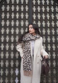 Manteau beige croisé, écharpe à motif léopard avec franges, tenant un sac à main marron foncé sur un fond métallique orné.
