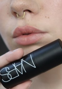 Gros plan sur des lèvres avec un anneau de septum en forme de cœur doré tenant un bâton de maquillage NARS noir près du visage.