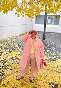 Άτομο με ροζ παλτό, καπέλο και γυαλιά στέκεται πάνω σε κίτρινα φθινοπωρινά φύλλα κοντά σε ένα κτίριο, κρατώντας ένα ροζ παγούρι.