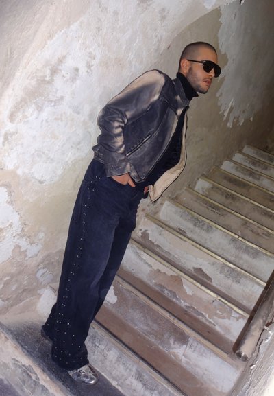 Chaqueta de cuero negra con detalles grises, cuello de tortuga negro, jeans oscuros con tachuelas en los laterales y zapatillas metálicas. De pie en una escalera.